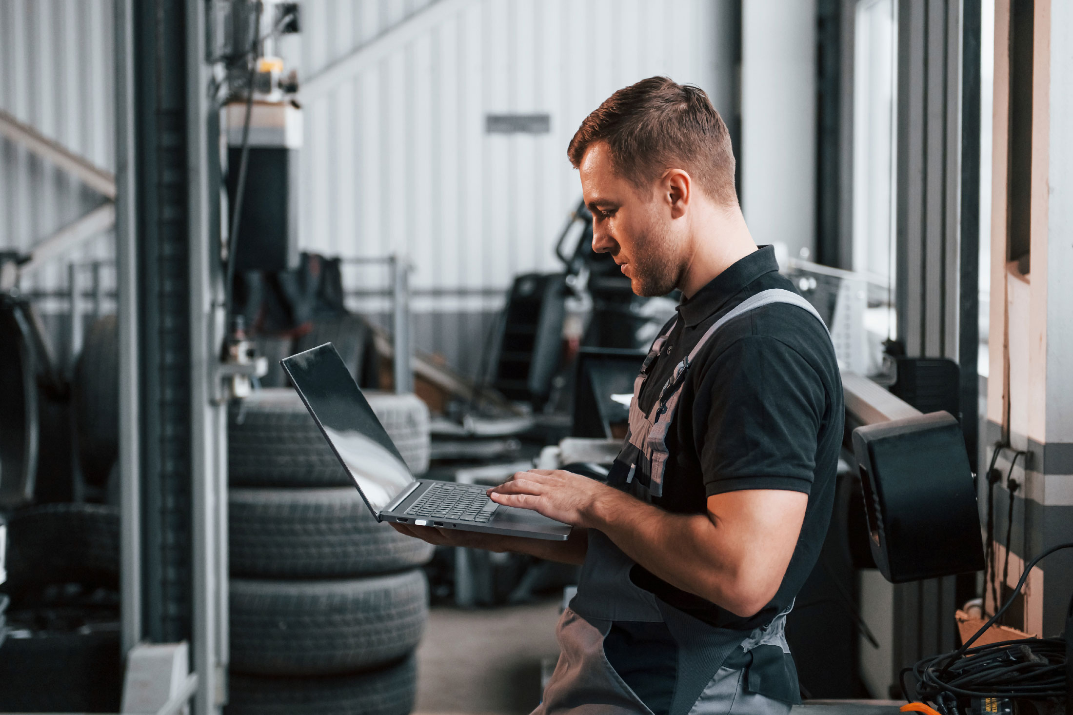 Young man in overalls uses laptop in a garage.