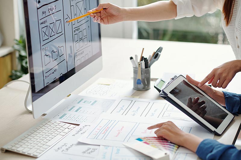 Two people analyze wireframe sketches on a computer screen and sheets of paper.