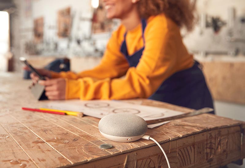 A young woman stands next to a Google Home device that sits on a table.