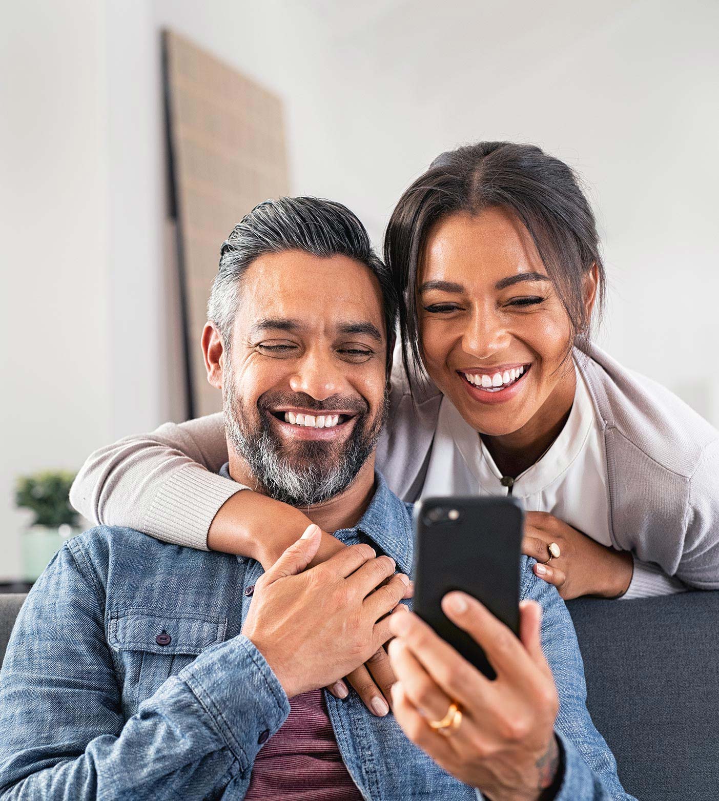 A woman drapes her arm over her husbands shoulders as he smiles at his phone.