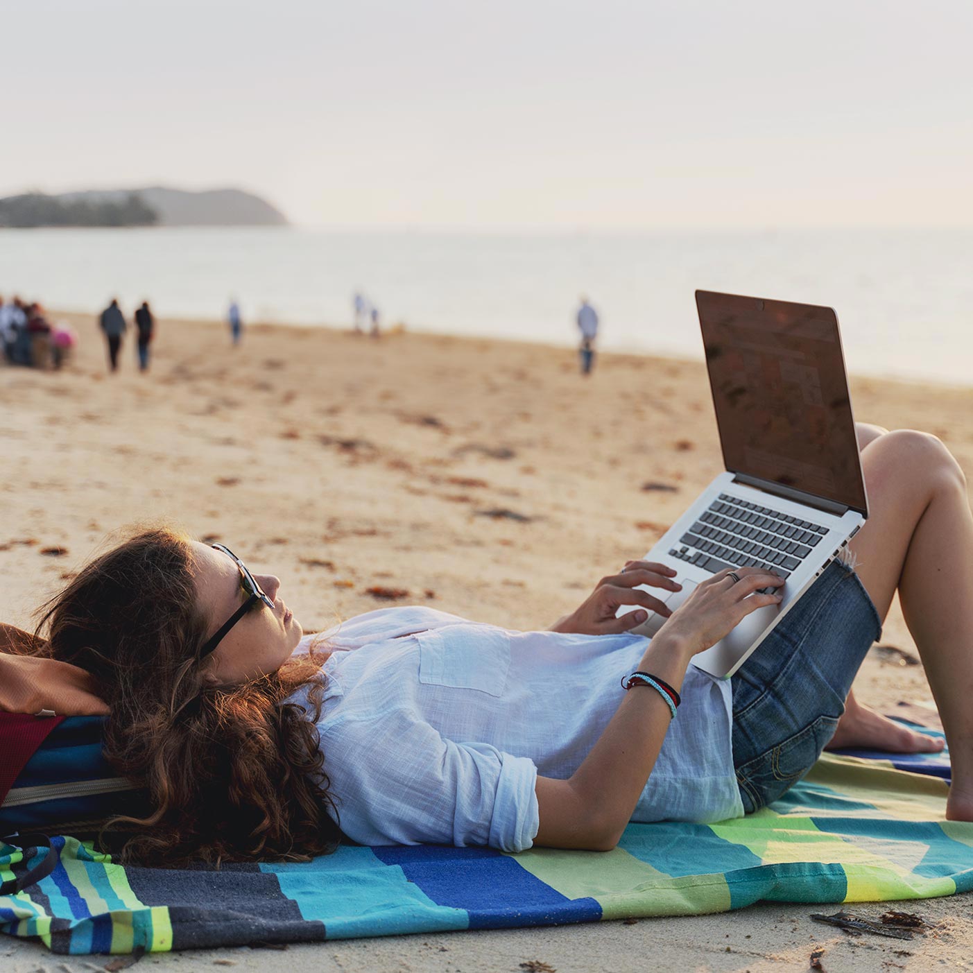A woman lies on the beach while working on her laptop that sits on her raised knees.