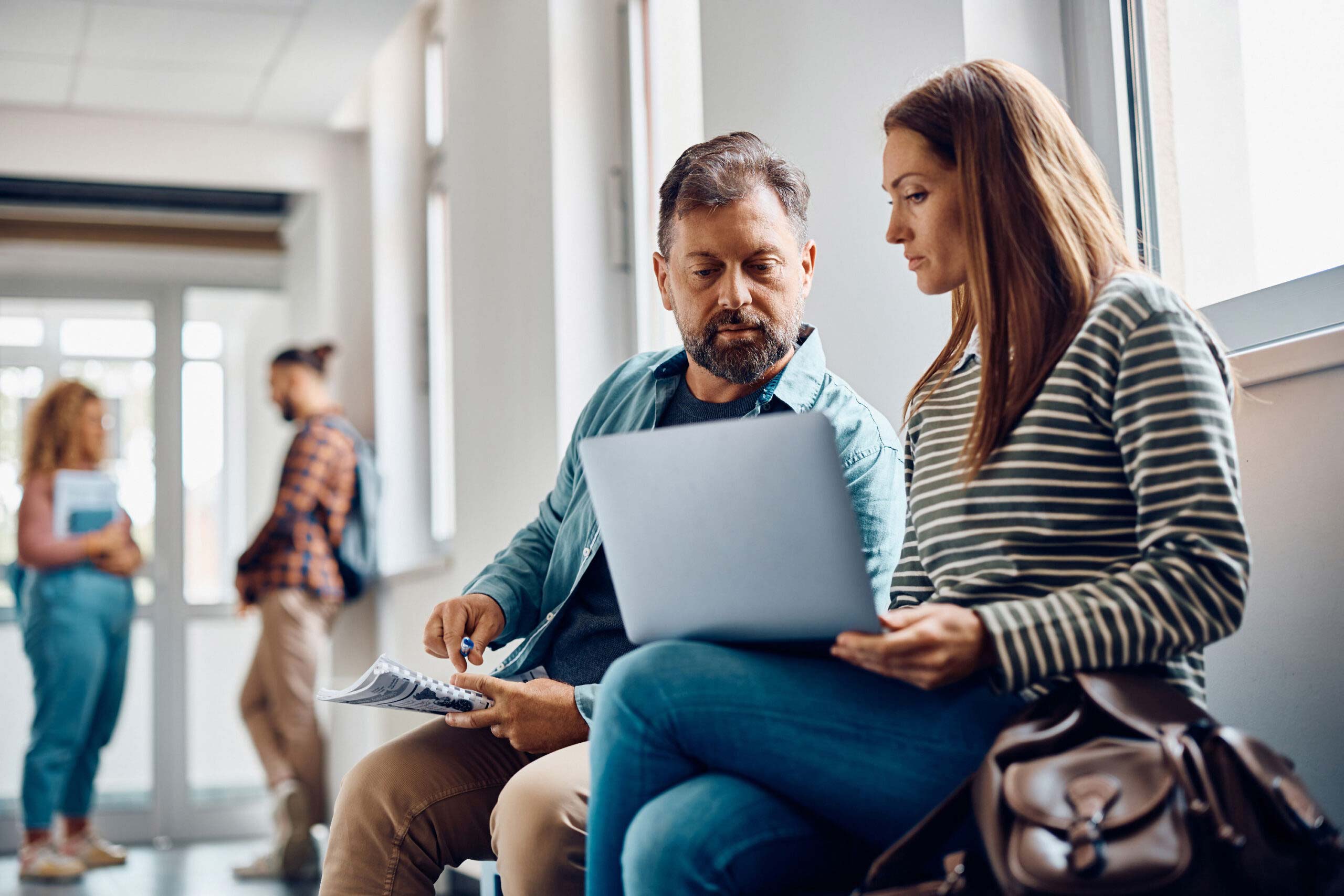 A man and woman sit together in a hallway discussing lessons on a laptop.