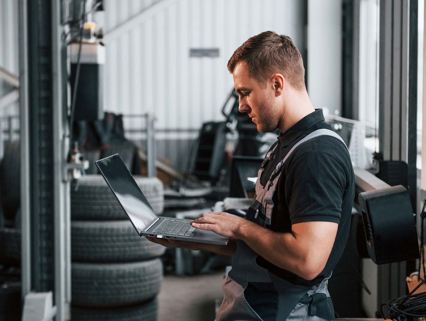 Young man in overalls uses laptop in a garage.