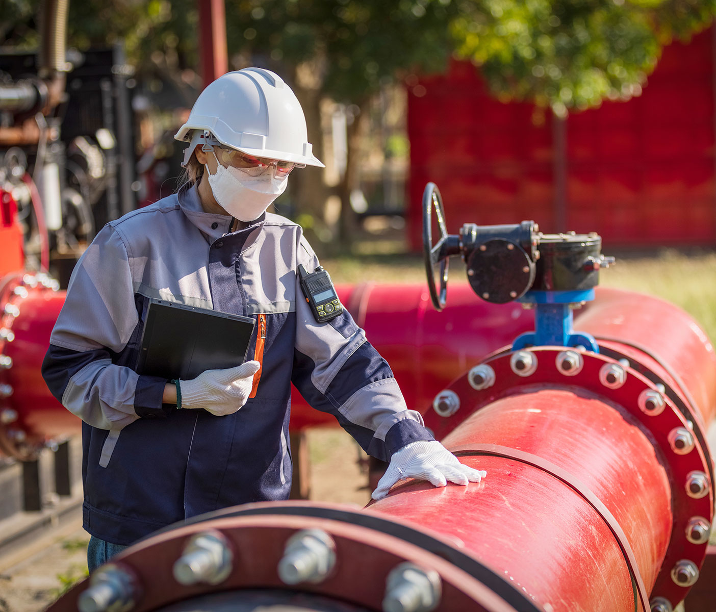 A worker in a hard hat examines a large pipeline while holding a tablet.
